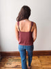 A woman viewed from behind wearing a burgundy camisole tank top with lace trim on the straps and blue jeans, standing on a wooden floor against a plain white wall.