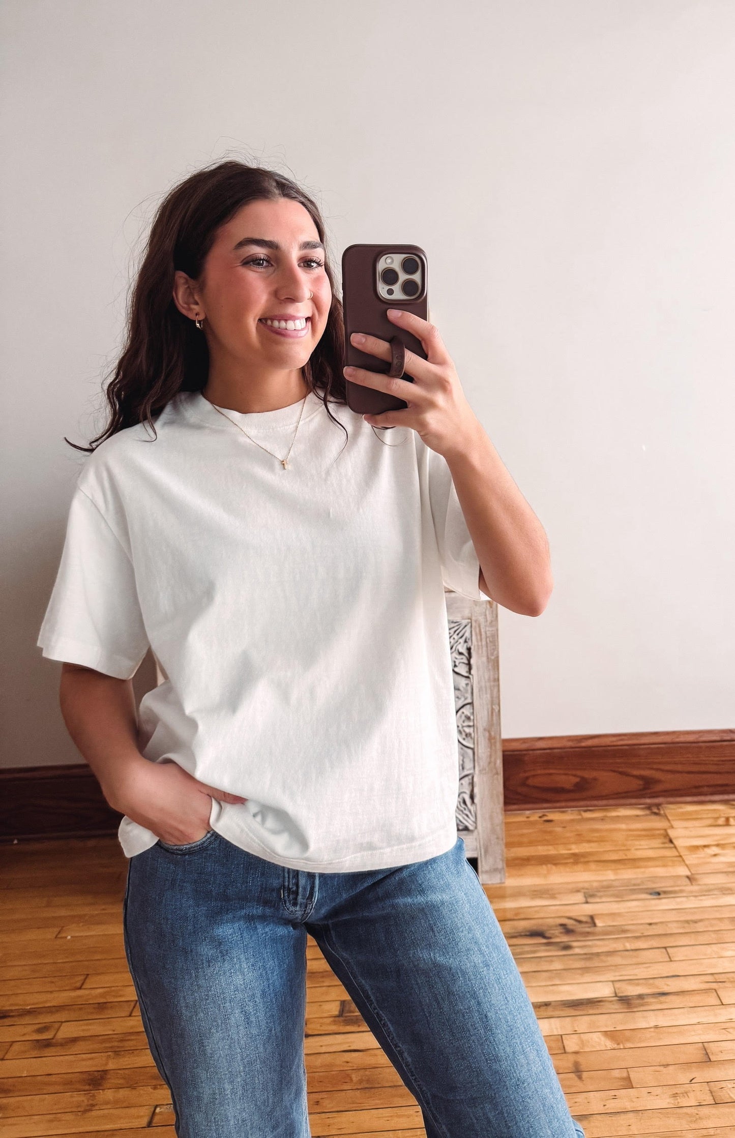 A woman taking a mirror selfie in a room with a light gray wall and wooden floor, wearing a white short-sleeve t-shirt and blue jeans.