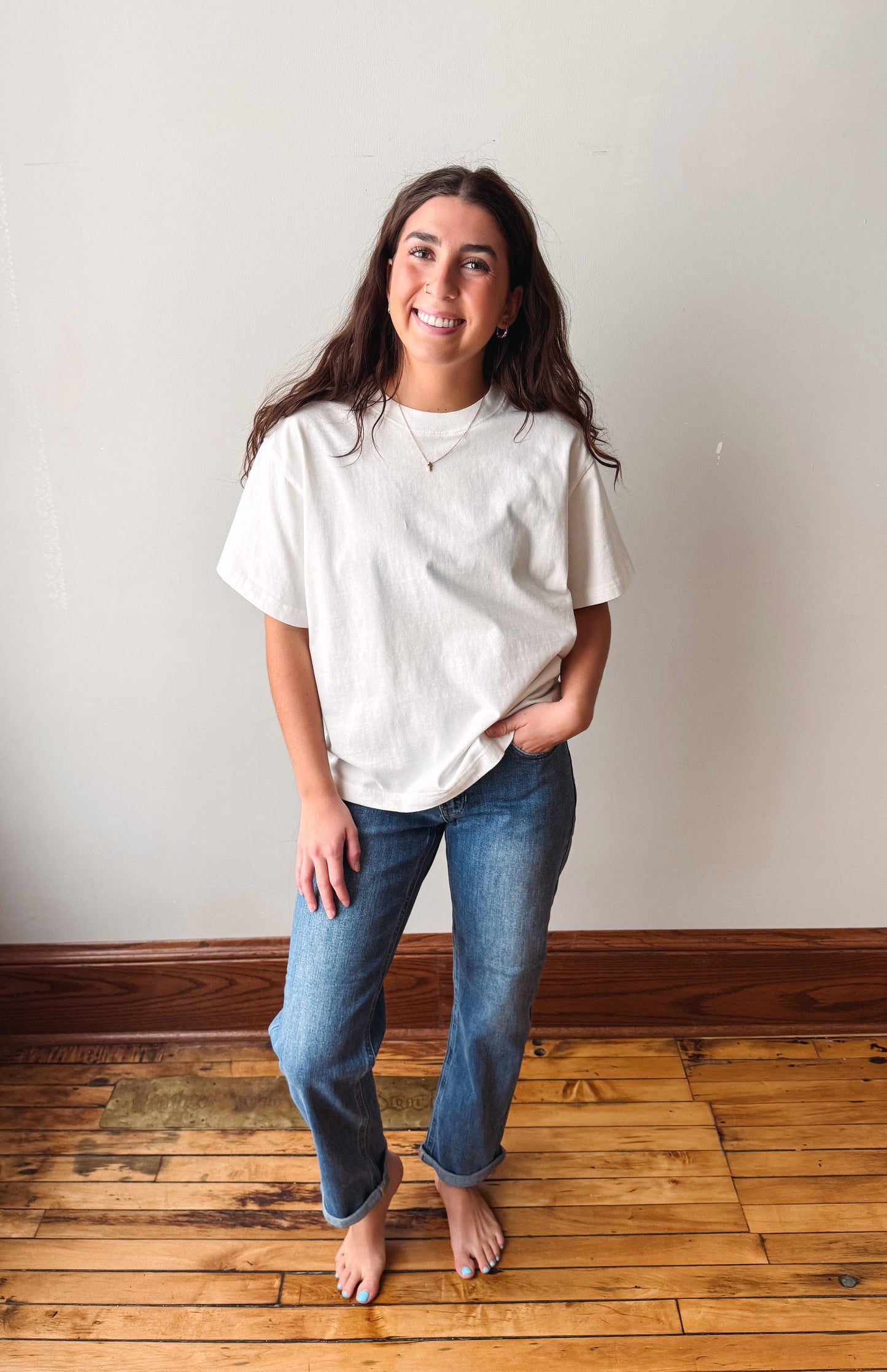 A woman stands barefoot on a wooden floor against a plain off-white wall, wearing an oversized off-white short-sleeve t-shirt and blue jeans.