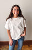 A smiling young woman with long brown hair stands indoors wearing an oversized off-white short-sleeve t-shirt and blue jeans against a plain light wall with a wooden baseboard and wood floor.
