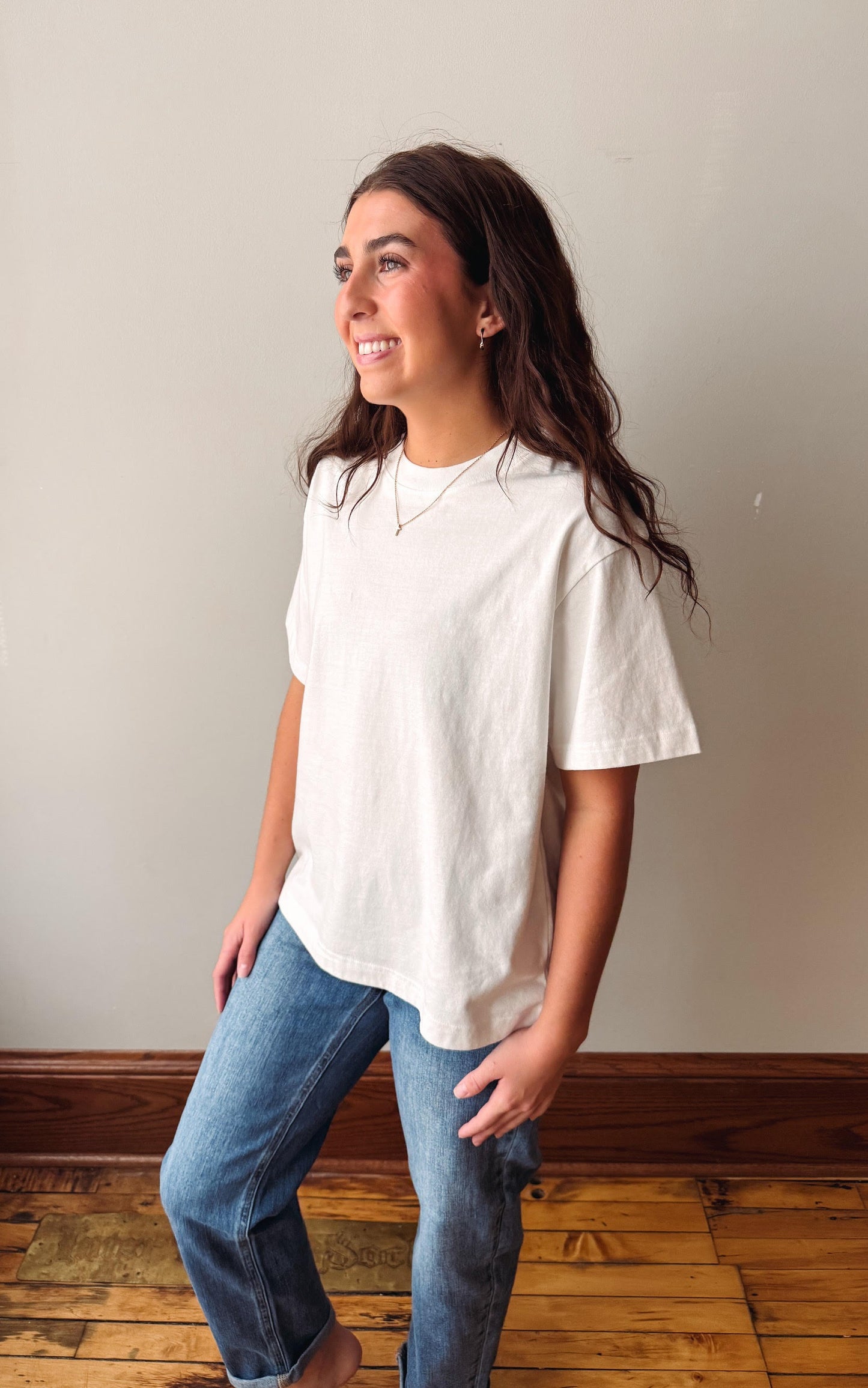 A woman wearing a loose-fitting, off-white short-sleeve t-shirt and blue jeans, standing in a room with a beige wall and wooden floor.
