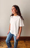A woman wearing a loose-fitting, off-white short-sleeve t-shirt and blue jeans, standing in a room with a beige wall and wooden floor.