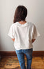 A person viewed from behind wearing an oversized white t-shirt and blue jeans, standing on a wooden floor against a plain off-white wall.