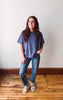 A woman modeling a loose-fitting slate blue t-shirt and blue jeans, standing on a wooden floor against a plain off-white wall.