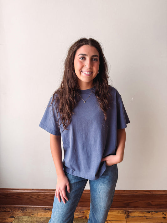 A woman with long brown hair smiling while wearing a slate blue short-sleeve t-shirt and blue jeans, standing against a plain off-white wall with a wooden baseboard and floor.