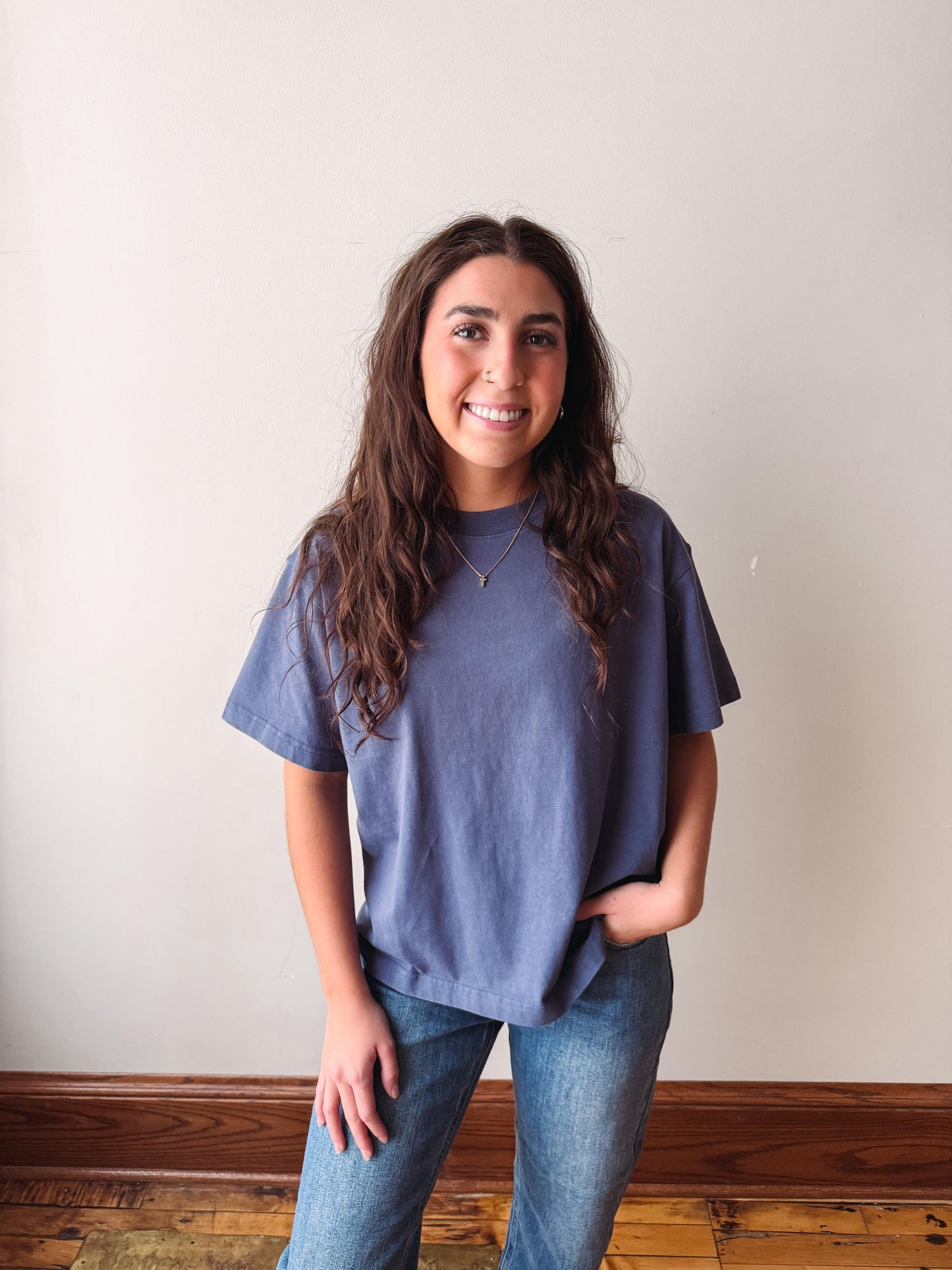 A woman with long brown hair smiling while wearing a slate blue short-sleeve t-shirt and blue jeans, standing against a plain off-white wall with a wooden baseboard and floor.
