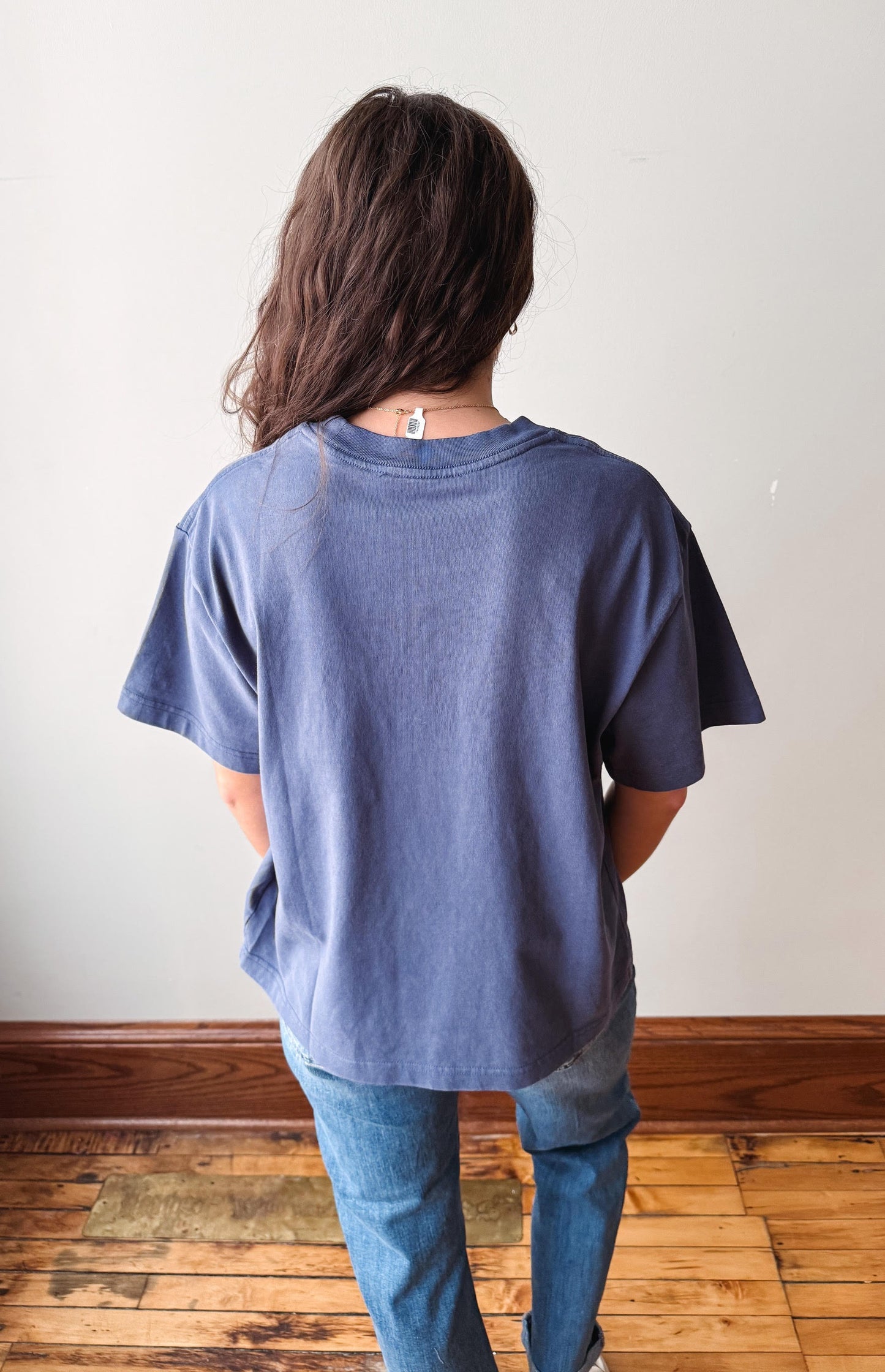 A person viewed from behind wearing an oversized faded blue t-shirt and blue jeans, standing on a wooden floor against a plain off-white wall.