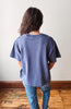 A person viewed from behind wearing an oversized faded blue t-shirt and blue jeans, standing on a wooden floor against a plain off-white wall.