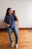 A woman takes a mirror selfie wearing a navy blue short-sleeve t-shirt, blue jeans, and white slide sandals in a room with a light gray wall and wooden floor.