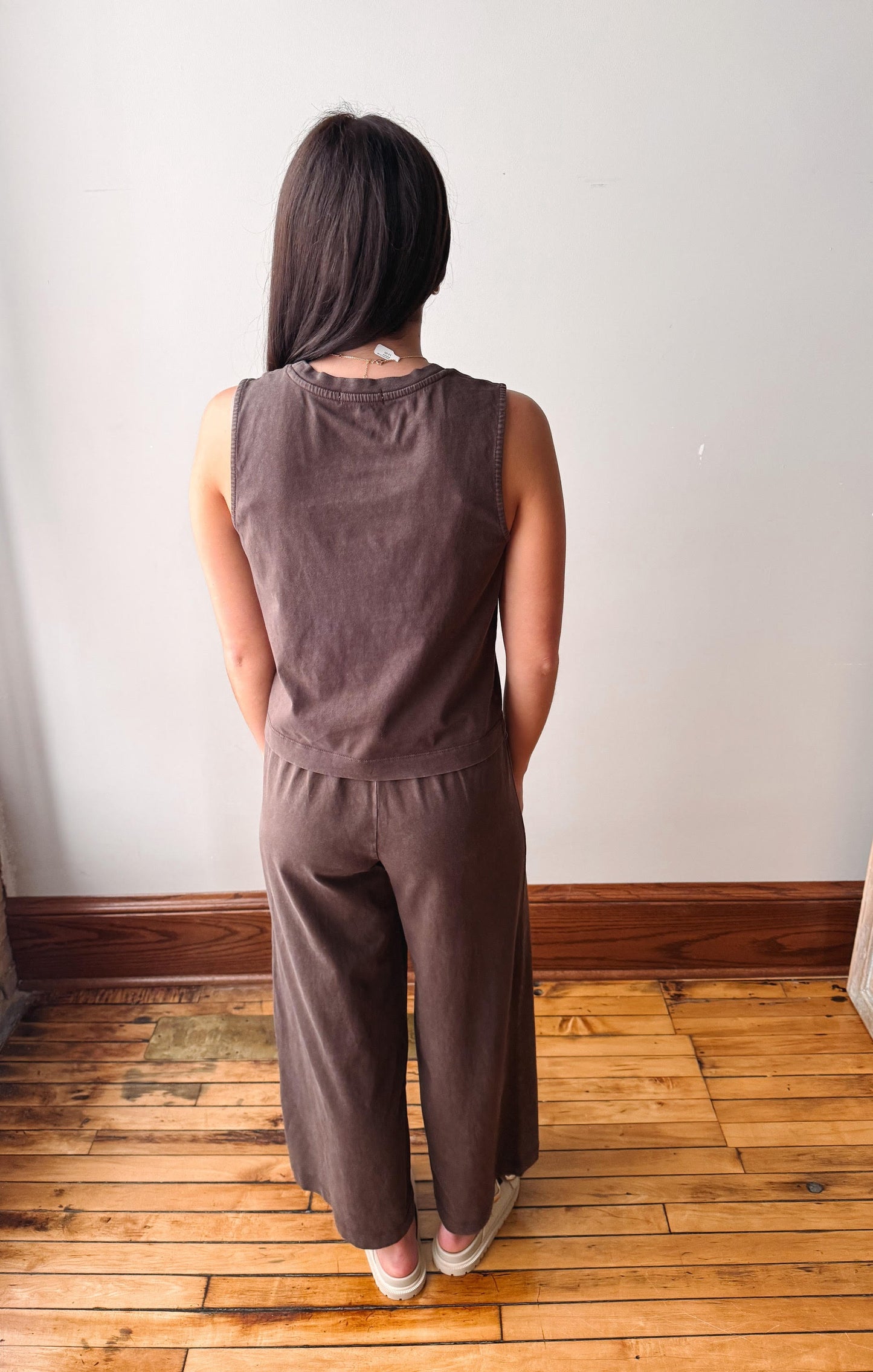 A woman viewed from behind wearing a matching sleeveless top and wide-leg pants set in a dark brown corduroy-like fabric, standing on a wooden floor against a plain white wall.