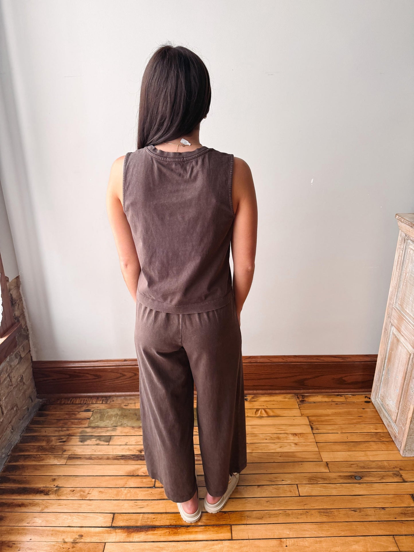 A woman viewed from behind wearing a matching sleeveless top and wide-leg pants set in a muted brown color, standing on a wooden floor against a plain wall.