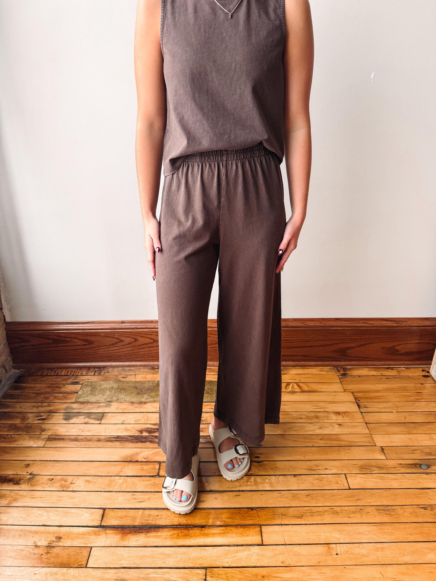 A person wearing a sleeveless taupe-brown top and matching wide-leg pants, standing on a wooden floor against a white wall.