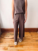 A person wearing a sleeveless taupe-brown top and matching wide-leg pants, standing on a wooden floor against a white wall.