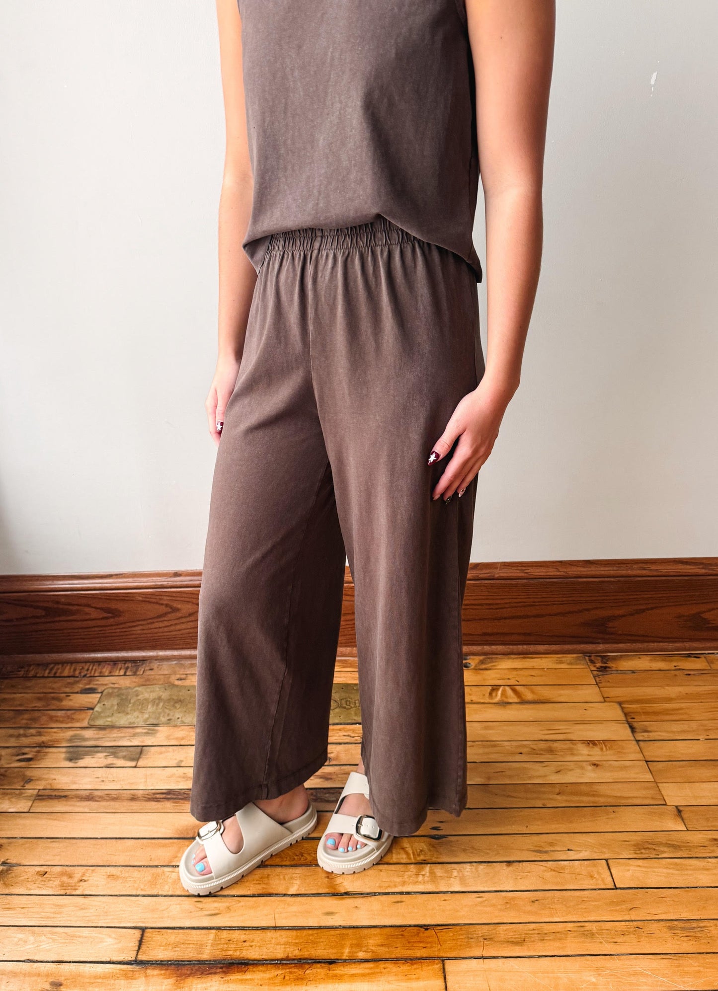 A person wearing a matching two-piece lounge set with a dark brown sleeveless top and wide-leg pants, standing on a wooden floor against a plain wall.