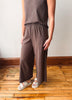 A person wearing a matching two-piece lounge set with a dark brown sleeveless top and wide-leg pants, standing on a wooden floor against a plain wall.