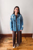 A woman stands in a room with a white wall and wooden floor, wearing a light blue denim jacket with white embroidered star and leaf patterns over a brown shirt and brown wide-leg pants.