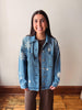 A woman wearing a light blue denim jacket with white embroidered star and leaf patterns, layered over a brown top and brown pants, standing against a plain off-white wall with wooden flooring.