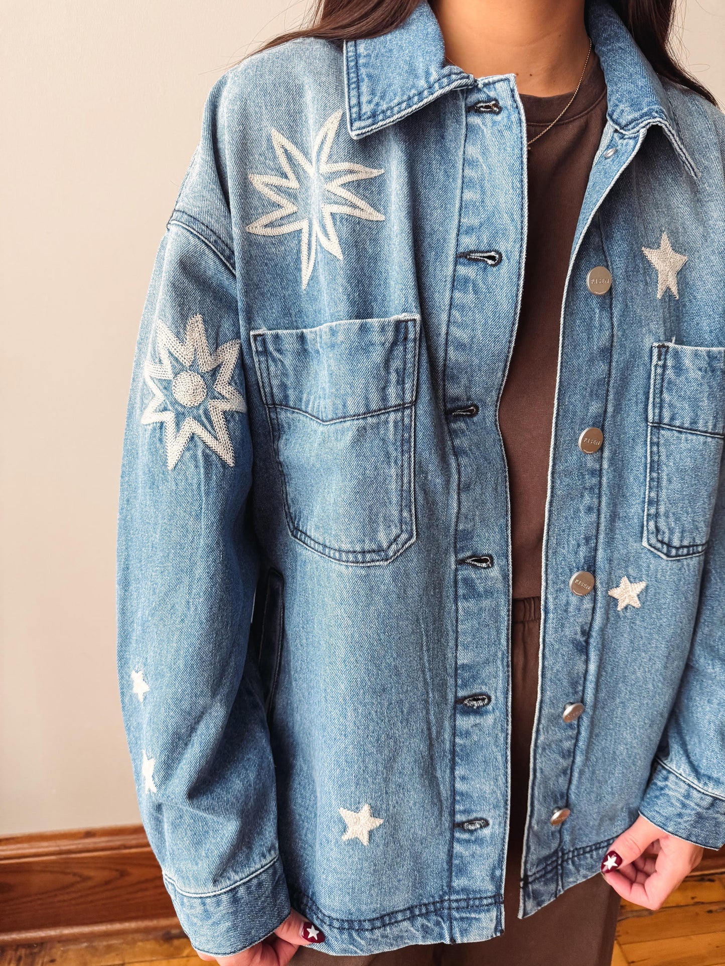 A person wearing a light blue denim jacket with white embroidered star and sunburst motifs, layered over a brown top. The photo is cropped from the neck down, set against a neutral beige wall and wooden floor.
