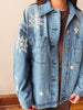 A person wearing a light blue denim jacket with white embroidered star and sunburst motifs, layered over a brown top. The photo is cropped from the neck down, set against a neutral beige wall and wooden floor.