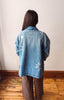 A person viewed from behind wearing an oversized light blue denim jacket with white star embroidery, paired with brown wide-leg pants, standing on a wooden floor against a plain off-white wall.