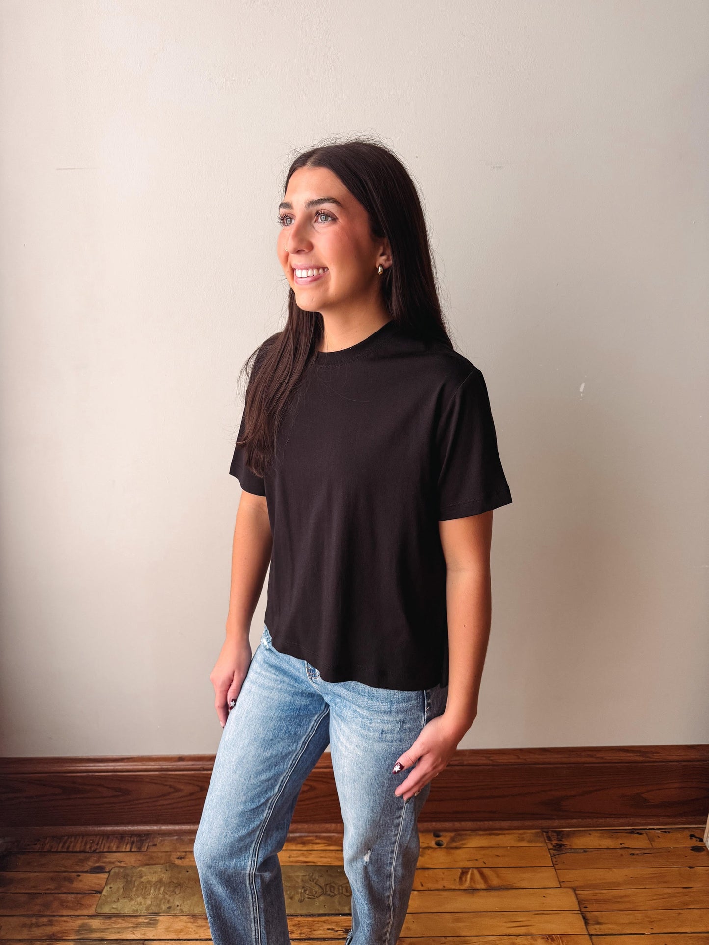 A woman with long dark hair stands in a room wearing a plain black short-sleeve t-shirt and light blue jeans, looking to the side against a beige wall and wooden floor.