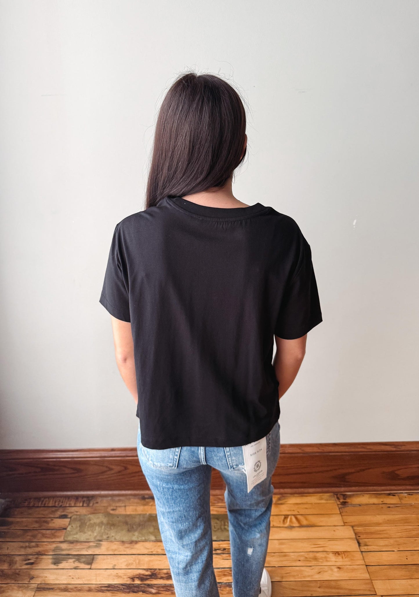 A woman viewed from behind wearing a plain black short-sleeve t-shirt and light blue jeans, standing on a wooden floor against a plain white wall.