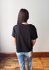 A woman viewed from behind wearing a plain black short-sleeve t-shirt and light blue jeans, standing on a wooden floor against a plain white wall.