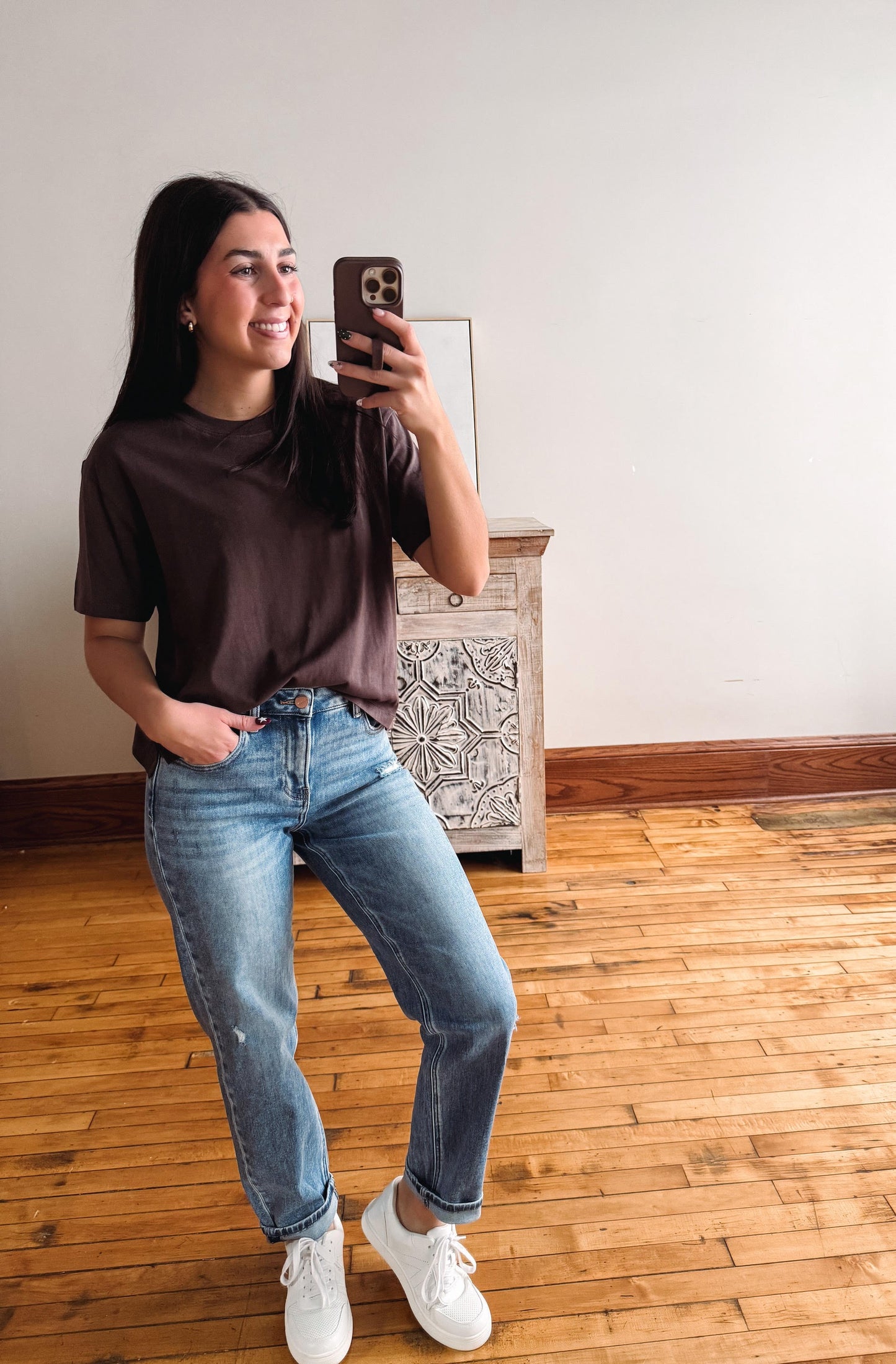 A woman taking a mirror selfie in a room with hardwood floors and a white wall, wearing a dark brown t-shirt, blue jeans, and white sneakers.