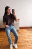 A woman taking a mirror selfie in a room with hardwood floors and a white wall, wearing a dark brown t-shirt, blue jeans, and white sneakers.
