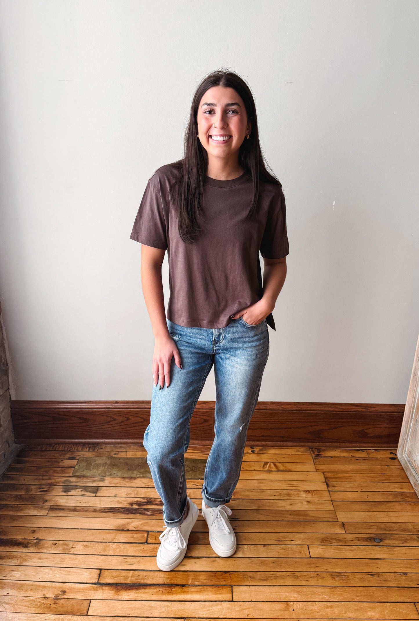 A woman stands in a room with a light gray wall and wooden floor, wearing a dark brown short-sleeve t-shirt, blue jeans, and white sneakers.