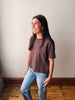A woman with long dark hair stands in a room wearing a brown short-sleeve crewneck t-shirt and light blue jeans, looking to the side.