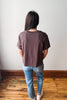 A person standing with their back to the camera, wearing a dark brown short-sleeve t-shirt, blue jeans, and white sneakers in an indoor setting with a white wall and wooden floor.