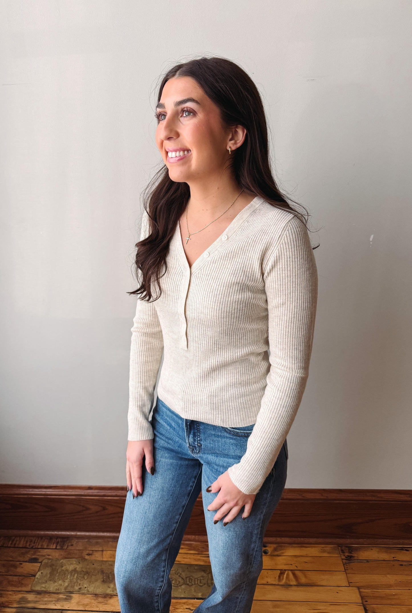A woman with long dark hair stands in a room wearing a ribbed cream-colored long-sleeve cardigan and blue jeans, looking off-camera.