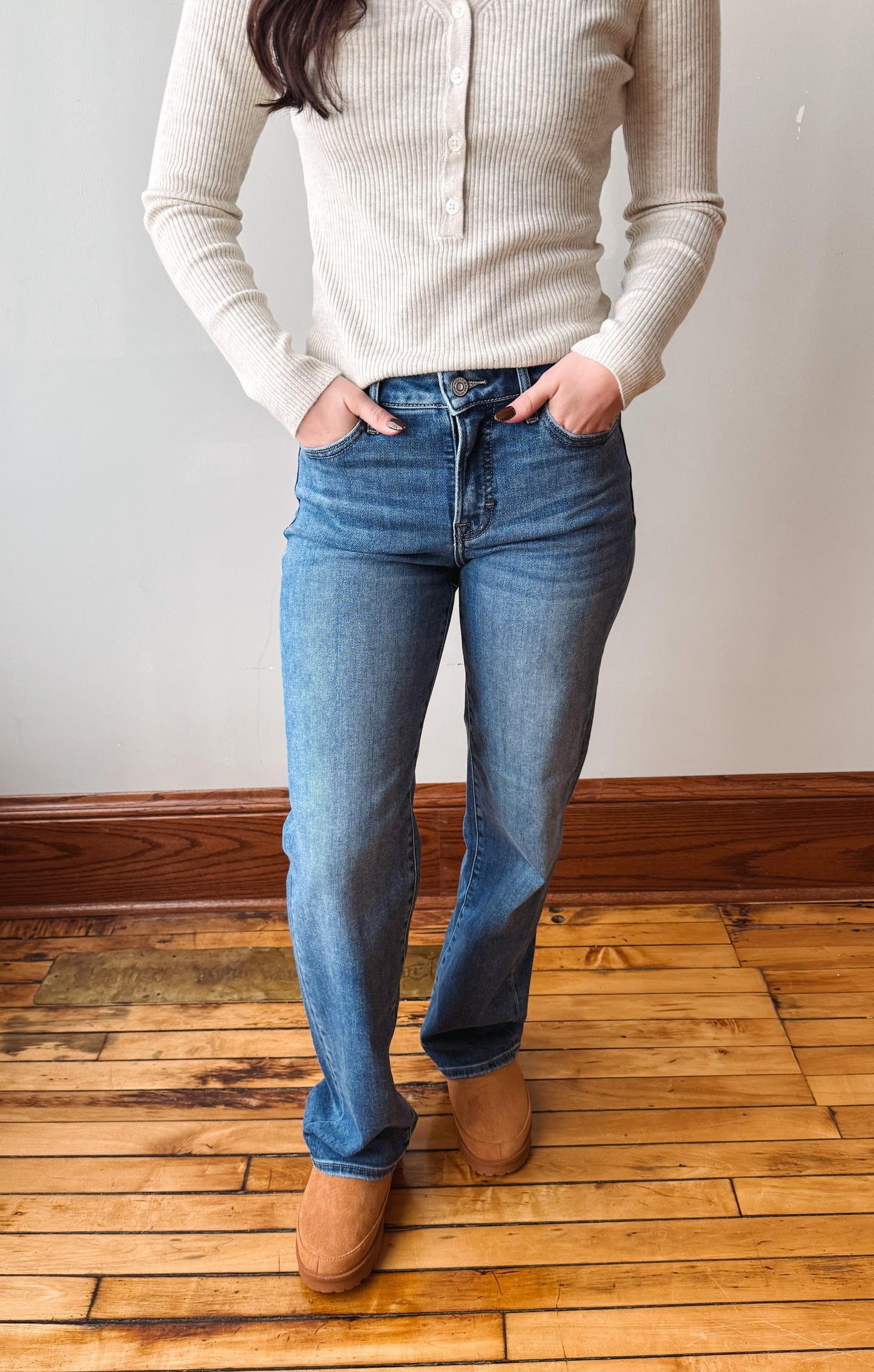 A person wearing blue straight-leg jeans, a cream ribbed knit sweater, and tan boots, standing on a wooden floor against a plain wall.