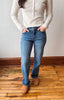 A person wearing blue straight-leg jeans, a cream ribbed knit sweater, and tan boots, standing on a wooden floor against a plain wall.