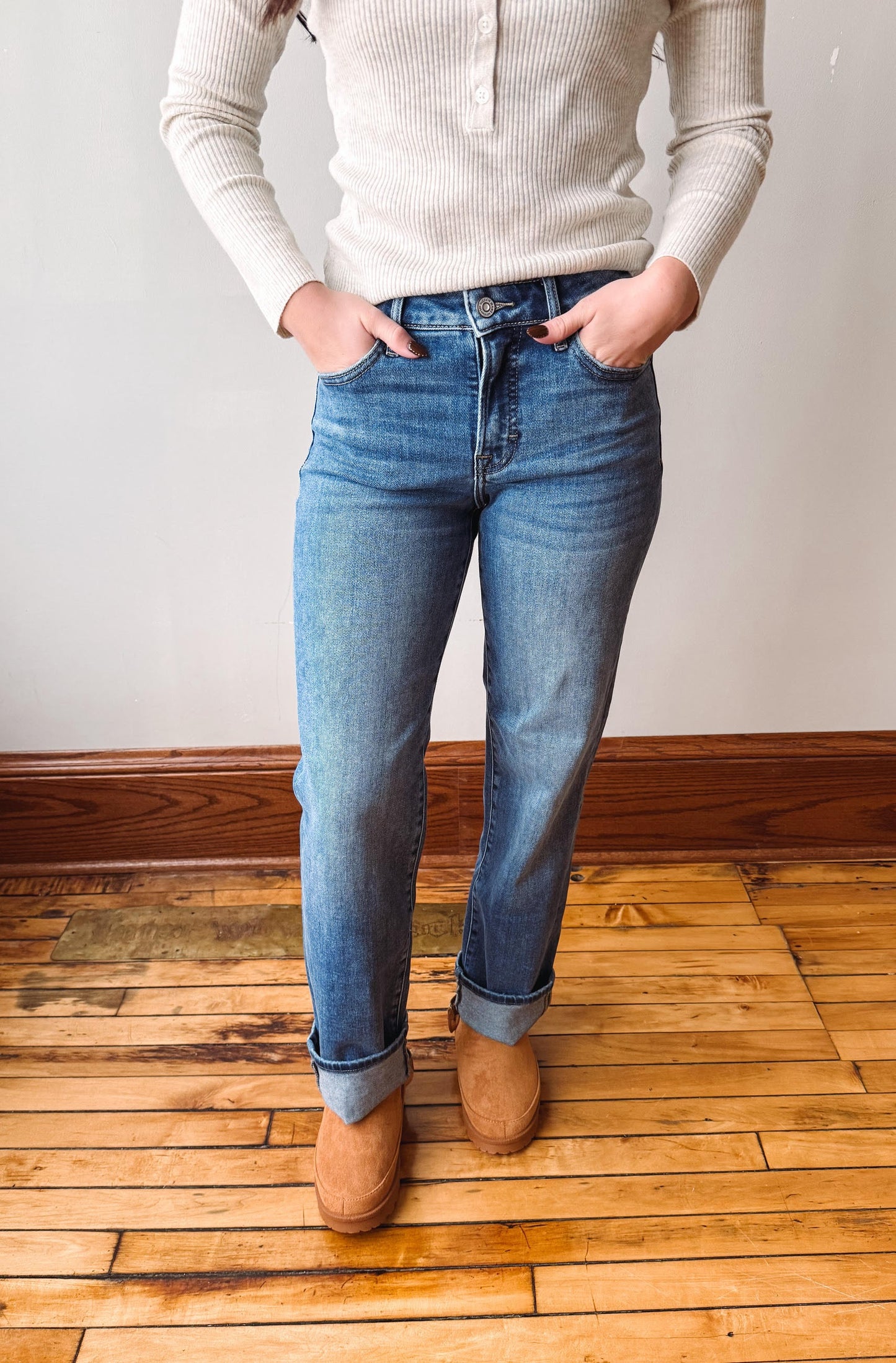 A person wearing medium-wash blue jeans with rolled cuffs, paired with a cream ribbed sweater and tan suede boots, standing on a wooden floor against a plain wall.
