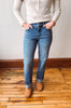 A person wearing medium-wash blue jeans with rolled cuffs, paired with a cream ribbed sweater and tan suede boots, standing on a wooden floor against a plain wall.