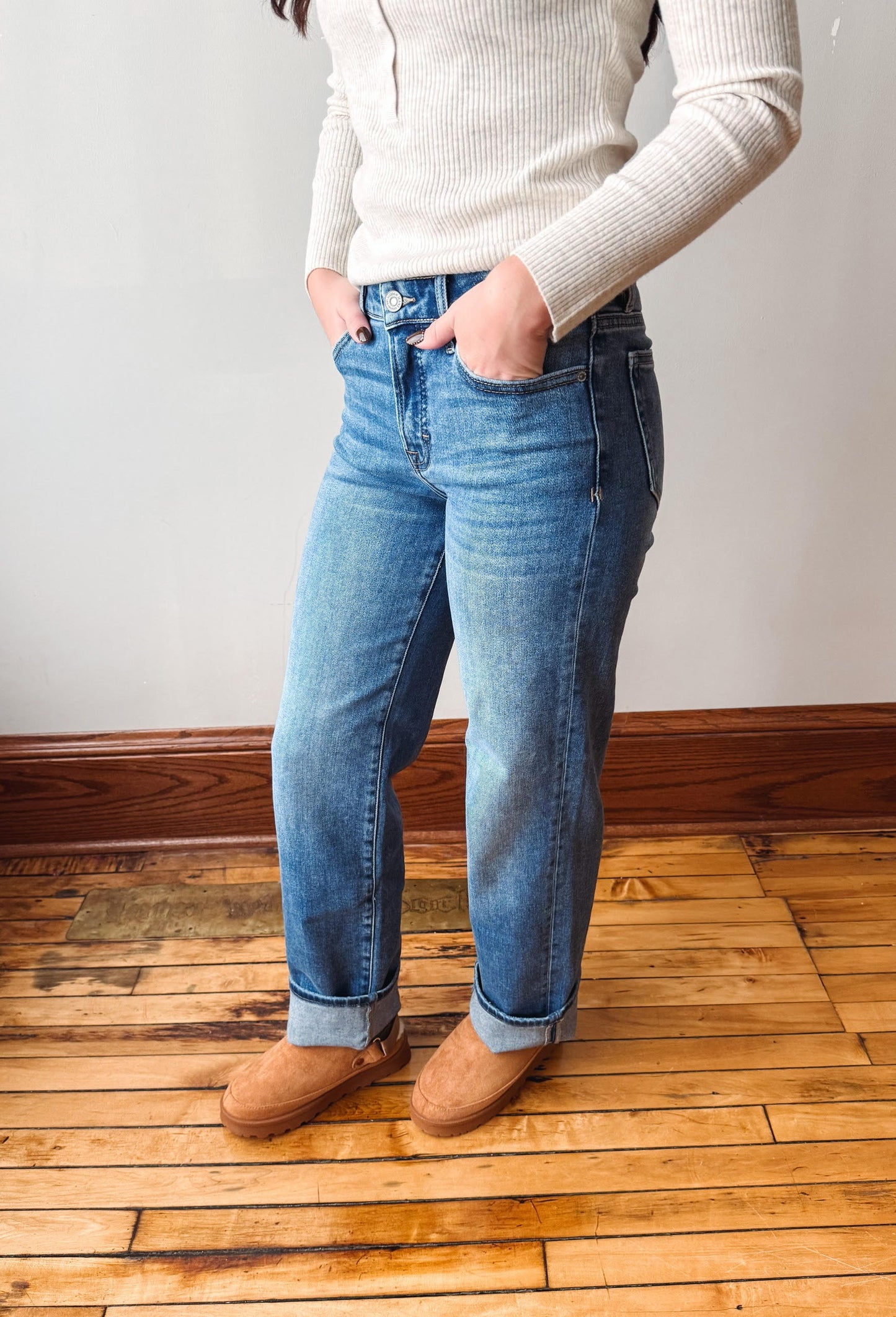 A person wearing blue straight-leg jeans with cuffed hems, paired with a cream ribbed long-sleeve top and brown suede boots, standing on a wooden floor against a white wall.