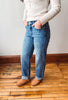 A person wearing blue straight-leg jeans with cuffed hems, paired with a cream ribbed long-sleeve top and brown suede boots, standing on a wooden floor against a white wall.