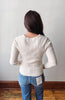 A woman viewed from behind wearing a cream ribbed long-sleeve top and blue jeans, standing against a plain light gray wall with a wooden baseboard and floor.