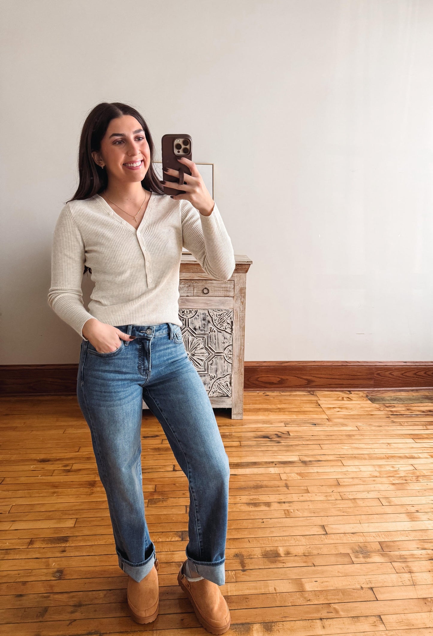 A woman takes a mirror selfie in a room with hardwood floors and a white wall, wearing a cream ribbed long-sleeve V-neck sweater, blue jeans, and tan slip-on shoes.