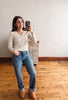 A woman takes a mirror selfie in a room with hardwood floors and a white wall, wearing a cream ribbed long-sleeve V-neck sweater, blue jeans, and tan slip-on shoes.