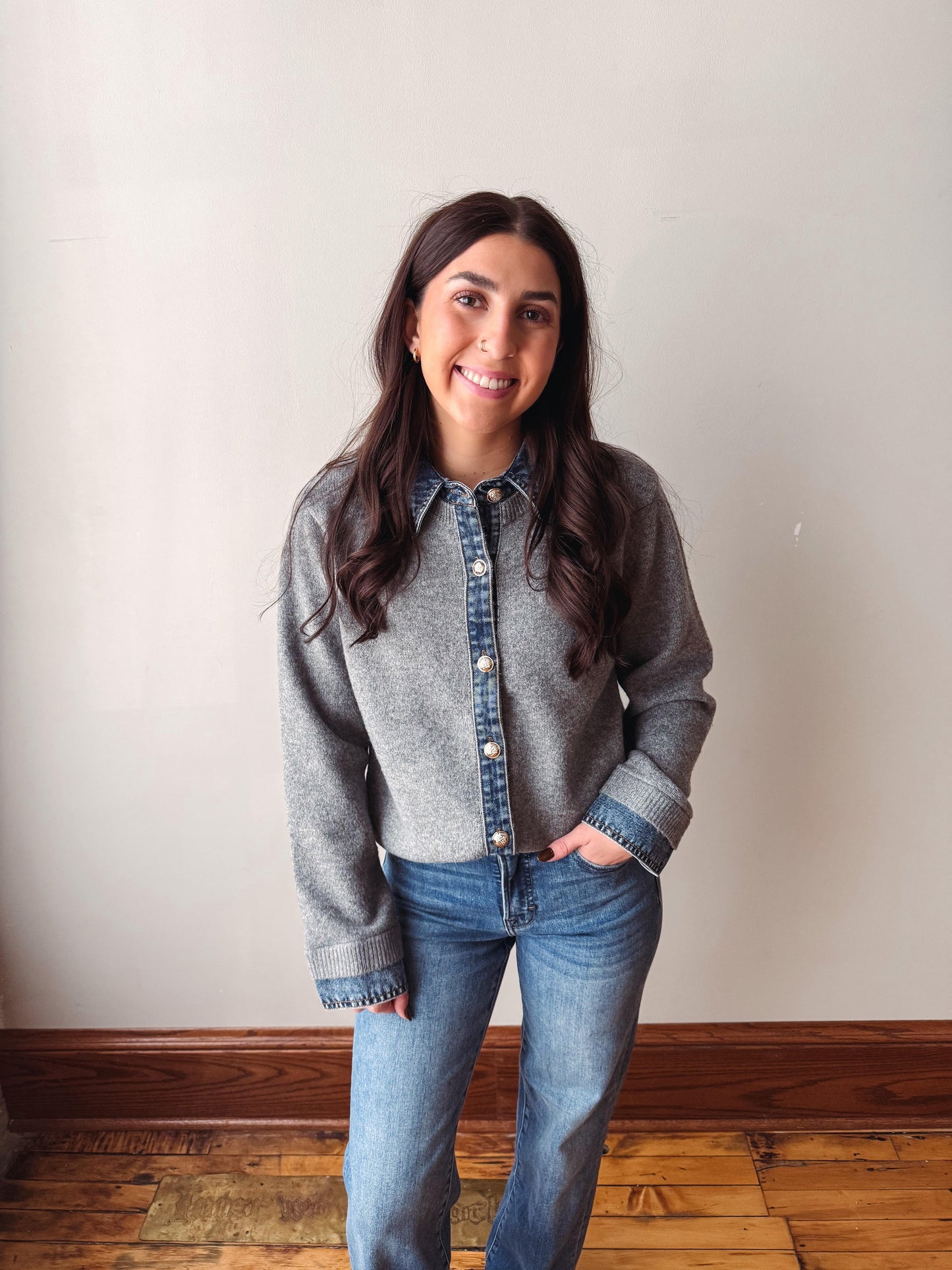 A woman with long dark hair stands indoors wearing a gray knit button-front cardigan layered over a blue denim shirt, paired with blue jeans, against a plain off-white wall with a wooden baseboard and floor.