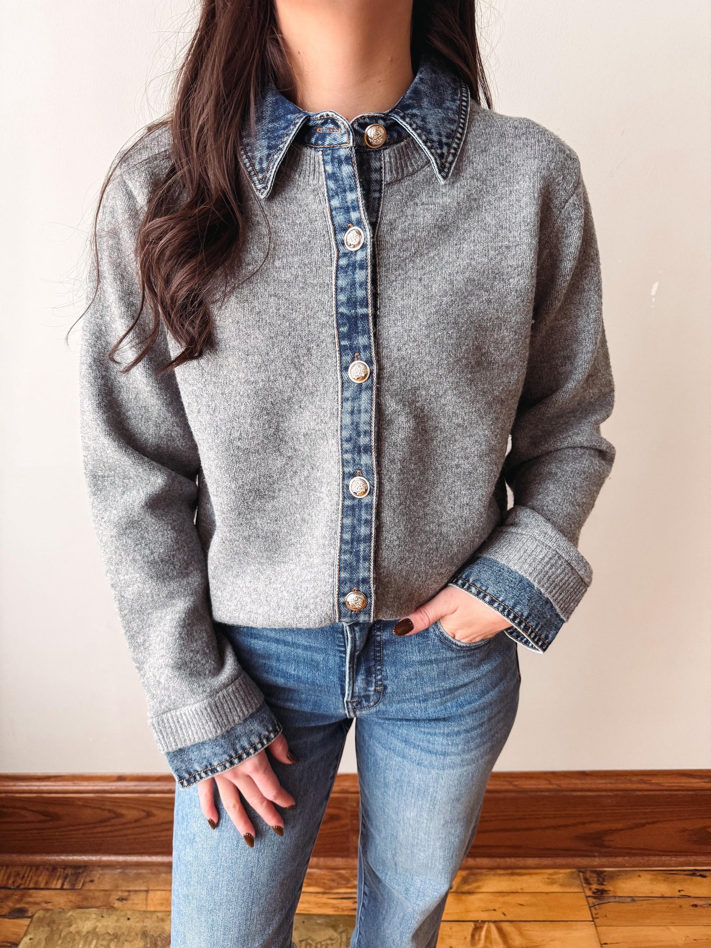 A person wearing a gray knit button-front cardigan with a denim collar and denim placket, paired with blue jeans, standing indoors against a plain light wall and wood floor.