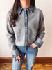 A person wearing a gray knit button-front cardigan with a denim collar and denim placket, paired with blue jeans, standing indoors against a plain light wall and wood floor.