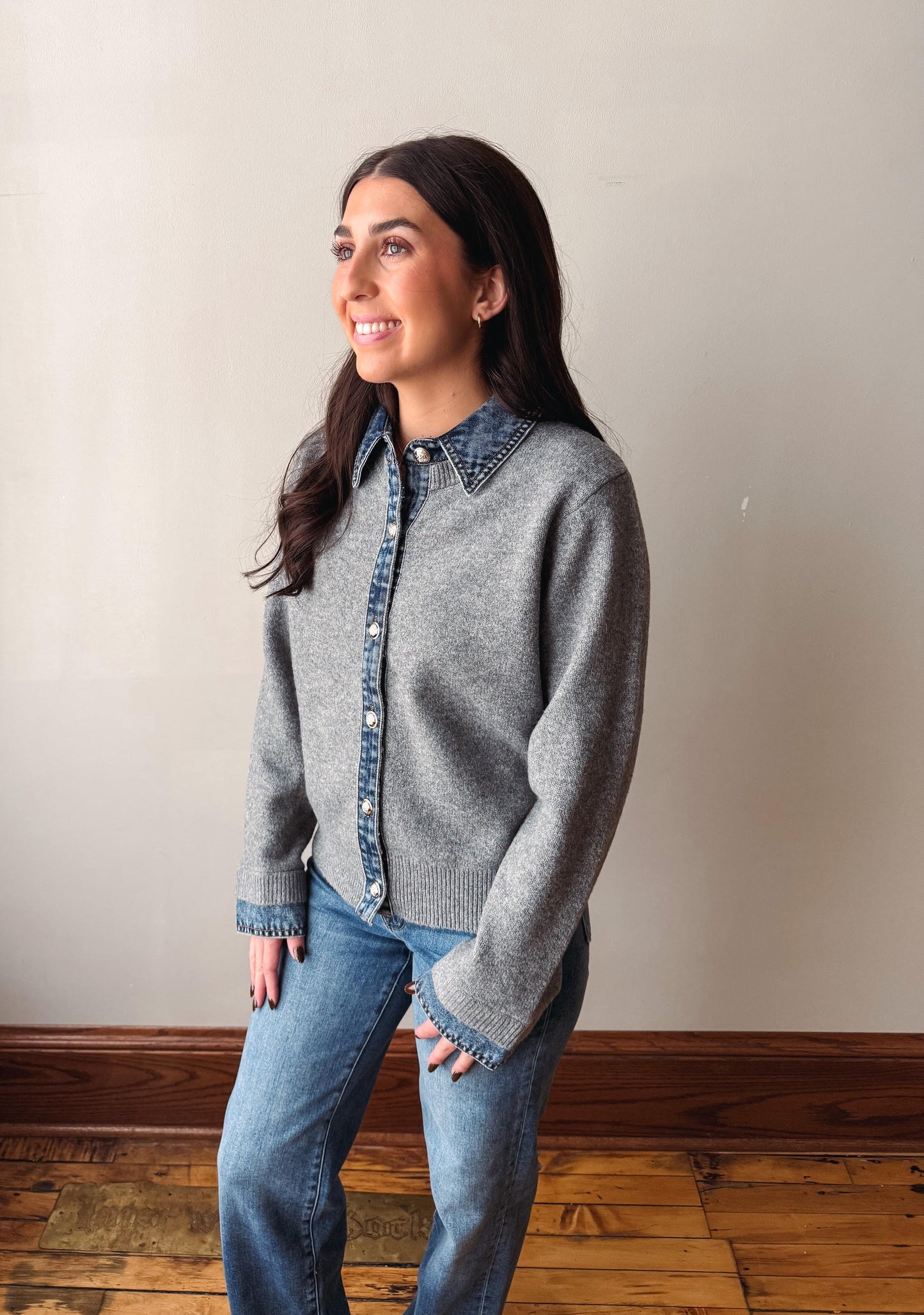 A woman wearing a gray knit cardigan with a denim button-down collar and front placket, paired with blue jeans, standing against a plain beige wall with a wooden floor.