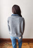A person viewed from behind wearing a heather gray knit sweater with a blue denim collar, paired with blue jeans, standing on a wooden floor against a plain off-white wall.