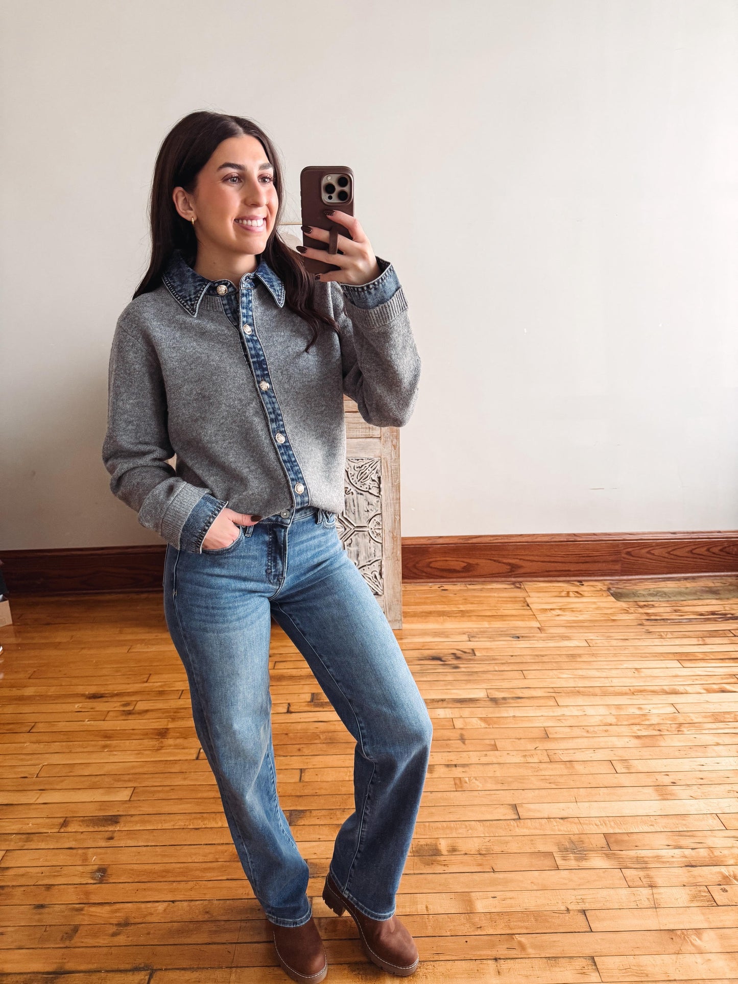 A woman takes a mirror selfie in a room with light wood floors and a white wall, wearing a gray knit button-up shirt with a blue denim collar and cuffs, blue jeans, and brown boots.