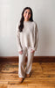 A woman modeling a matching cream cable-knit sweater and wide-leg knit pants set, standing on a wooden floor against a plain off-white wall.