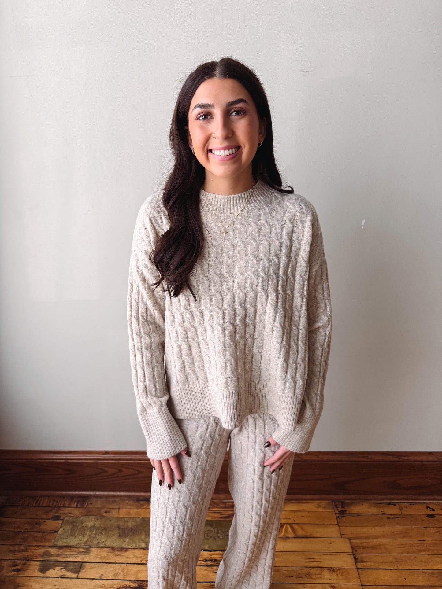 A woman wearing a matching cream cable-knit sweater and pants set, standing in front of a plain off-white wall with a wooden baseboard and hardwood floor.
